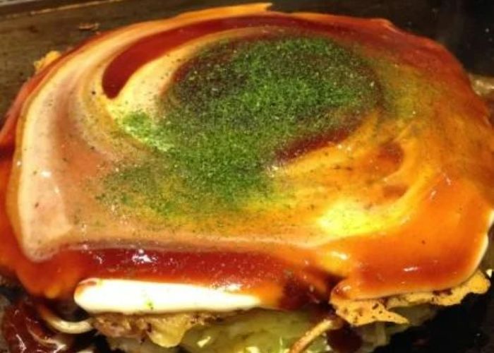 A freshly cooked okonomiyaki at Tsuruhashi Fugetsu Odaiba, showing a saucy and herb-covered fried egg on top of cabbage and batter.
